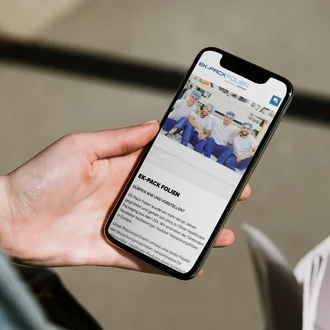 Hand hält Smartphone mit geöffneter Website von EK-Pack Folien – zu sehen sind vier junge Männer in Arbeitskleidung, die lächelnd posieren. Auf dem Bildschirm wird die Unternehmensvorstellung von EK-Pack Folien angezeigt