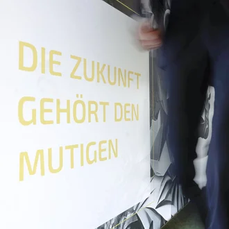 Person geht dynamisch an Wand mit motivierendem Schriftzug ‚Die Zukunft gehört den Mutigen‘ im Denkströme Büro vorbei