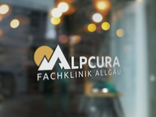 Glasfront mit Logo-Fensterbeklebung der Alpcura Fachklinik Allgäu vor unscharfem Innenraum mit warmen Lichtreflexionen im Hintergrund.
