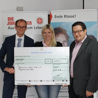 Spendenübergabe an die Johanniter-Unfall-Hilfe 2018: Drei Personen – zwei Männer im Anzug und eine Frau mit langem blondem Haar – halten einen großen symbolischen Scheck über 400 Euro vor einem Johanniter-Banner in Kempten