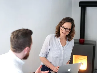 Zwei Denkströme Kollegen im Gespräch mit Laptop bei Besprechung mit Kaminofen im Hintergrund – kreative Zusammenarbeit in gemütlicher Arbeitsumgebung