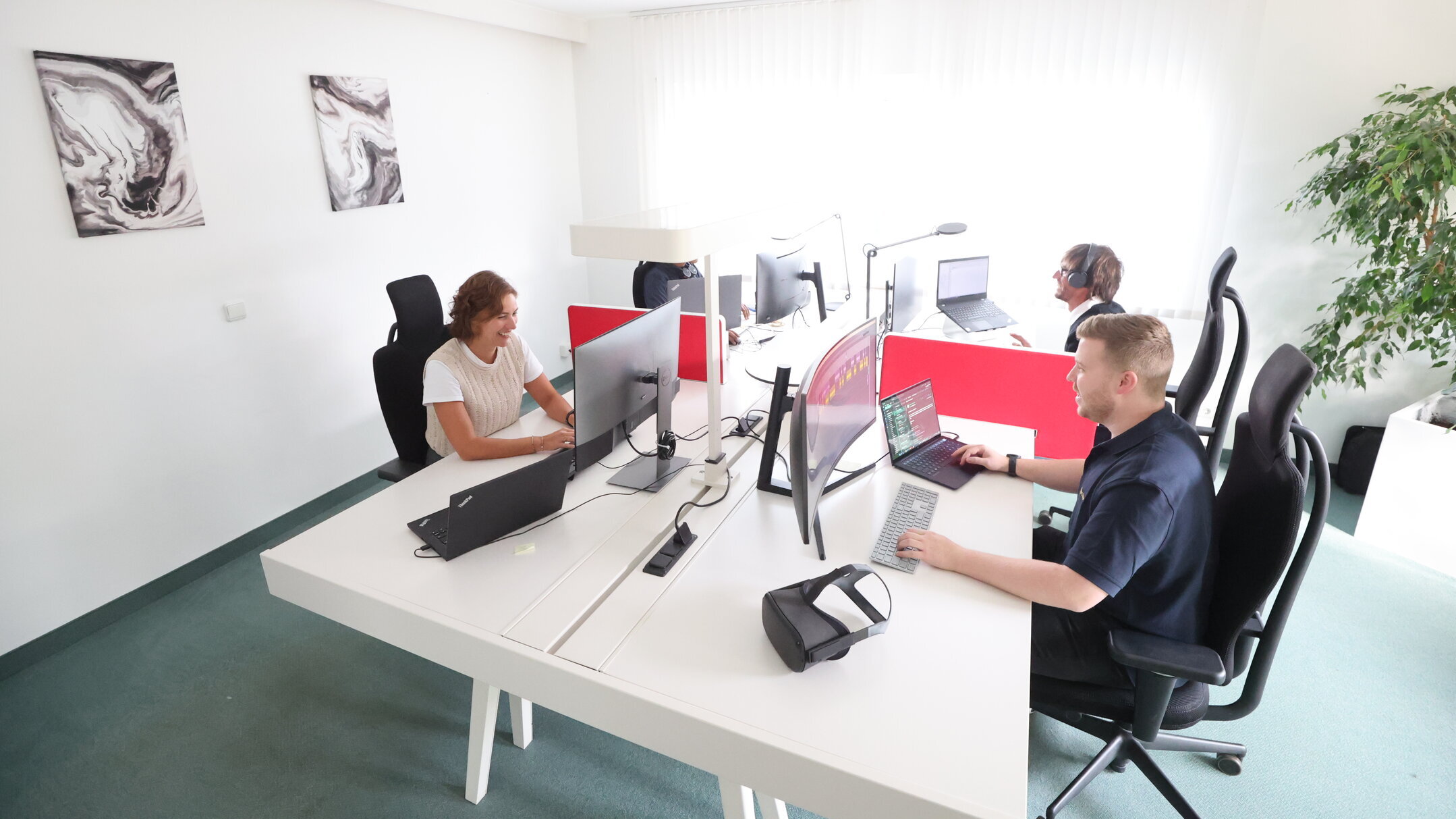 Denkströme Team arbeitet im hellen Großraumbüro an mehreren Monitoren und Laptops, getrennt durch rote Sichtschutzwände – Arbeitsplatzsituation für Website-Relaunch und digitale Projekte.