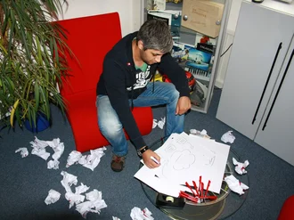 Kreativer Mitarbeiter sitzt in modernem Büro auf rotem Sessel und skizziert Konzeptideen – umgeben von zerknülltem Papier und Brainstorming-Notizen