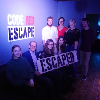 Gruppenfoto eines erfolgreichen Escape-Room-Abenteuers im CODE RED ESCAPE: Acht Personen, lachend und entspannt, halten ein Schild mit der Aufschrift ‚ESCAPED‘ – Teambuilding-Event 2018 in stimmungsvoll beleuchtetem Raum