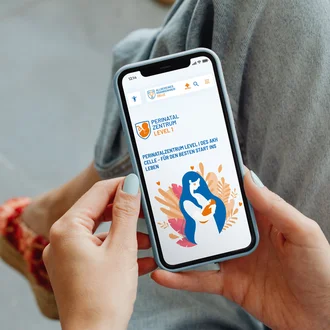 Frau hält Smartphone mit geöffneter Website des Perinatalzentrums Level 1 am Allgemeinen Krankenhaus Celle – digitale Startseite mit Illustration einer Mutter und Baby für den besten Start ins Leben