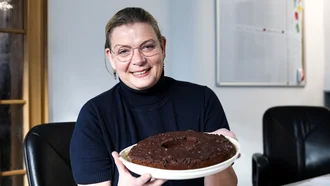Freundlich lächelnde Denkströme Mitarbeiterin im Büro hält einen Schokoladenkuchen in den Händen