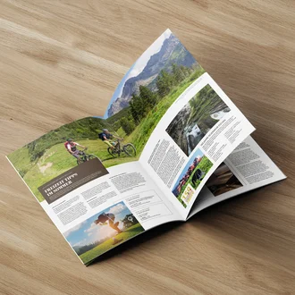 Sommerjournal mit Freizeit-Tipps, Radfahrern in den Alpen und Ausflugszielen in aufgeschlagener Magazinansicht auf Holzoberfläche