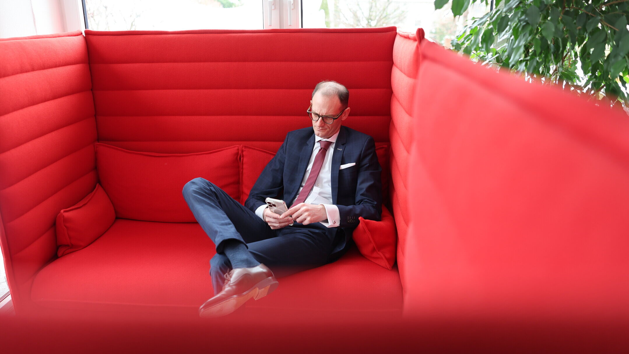 Denkströme Geschäftsführer im Anzug sitzt entspannt in moderner roter Lounge-Sitznische und nutzt Smartphone – Rückzugsort für konzentriertes Arbeiten im Büro.