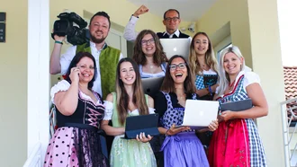 Team der Denkströme in Tracht mit Laptops und Kamera auf einer Treppe – gelebte Unternehmenskultur trifft digitale Kreativität in traditioneller Kleidung