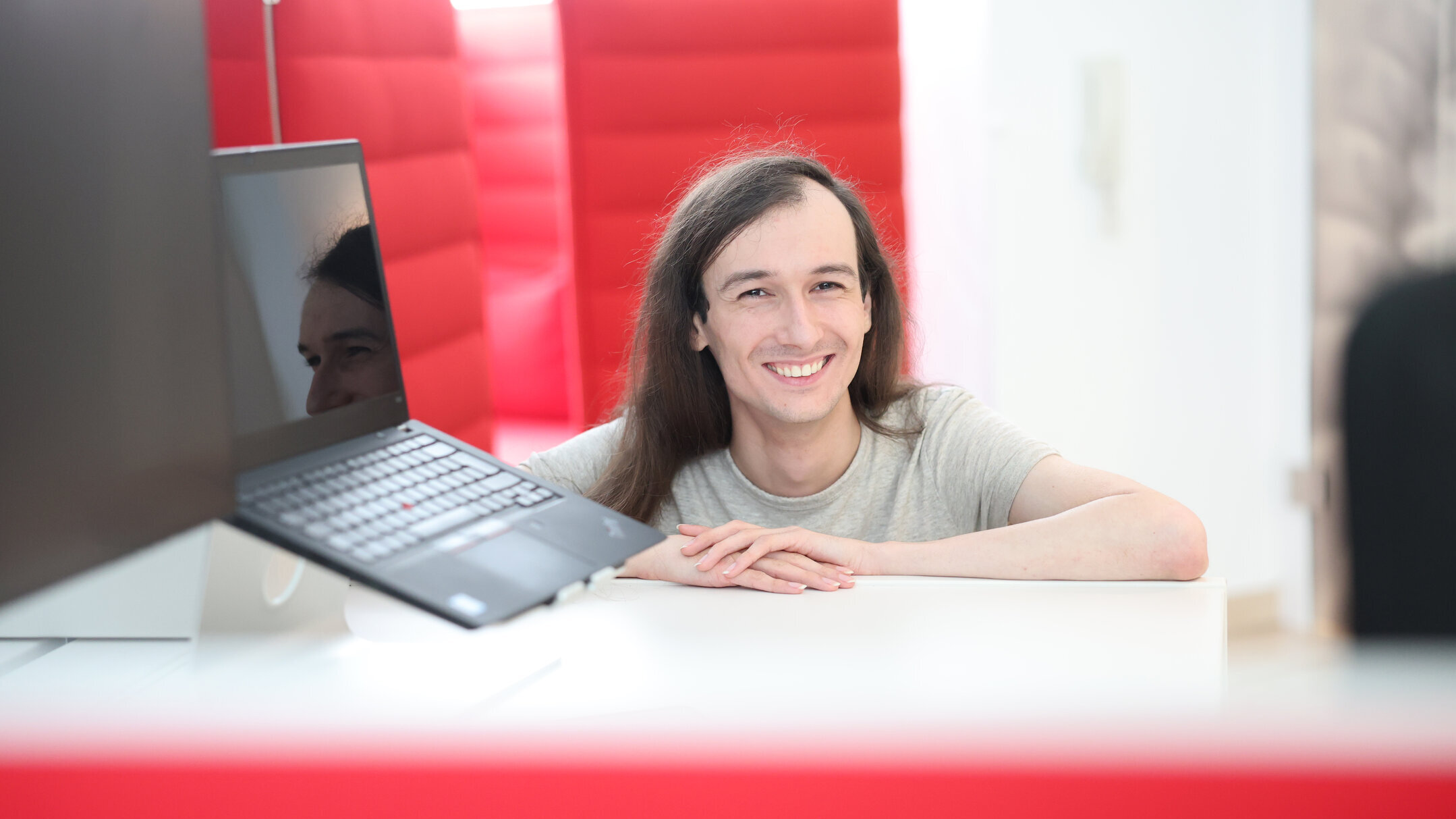 Mitarbeiter aus der Grafikabteilung von Denkströme lächelt im Büro hinter einem Schreibtisch; Laptop und Monitor stehen im Vordergrund, rote Akustikelemente im Hintergrund.