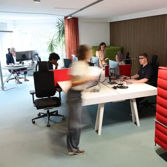 Offenes Büro bei Denkströme in Kempten: Mitarbeitende arbeiten an Schreibtischen mit Monitoren, eine Person bewegt sich durch den Raum; moderne Arbeitsplätze mit roten Akustikelementen.