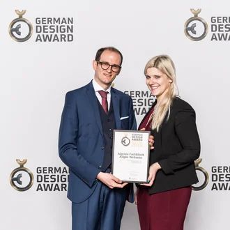 Preisträger des German Design Award 2019 – zwei Agenturvertreter mit Urkunde für die Webseite der Alpcura Fachklinik Allgäu vor Award-Backdrop, elegante Businesskleidung, professionelle Auszeichnung