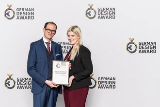 Preisträger des German Design Award 2019 – zwei Agenturvertreter mit Urkunde für die Webseite der Alpcura Fachklinik Allgäu vor Award-Backdrop, elegante Businesskleidung, professionelle Auszeichnung