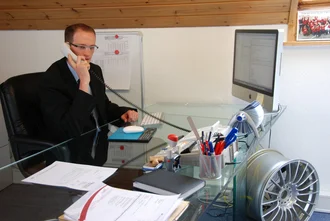 Büroszene aus dem Jahr 2008: Mann im Anzug telefoniert am Schreibtisch mit iMac und Glasplatte, modernes Büro mit Felge als Designelement
