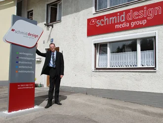 Inhaber Fabian Schmid im Anzug vor dem Gebäude der schmid design media group mit großem Unternehmensschild für Werbeagentur, Internetagentur, Drucktechnik und Eventmarketing