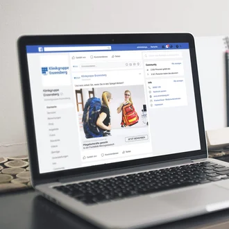 Facebook-Post der Klinikgruppe Enzensberg auf Laptopbildschirm zeigt Pflegekampagne mit zwei jungen Frauen und Rucksäcken im Dialogmotiv.