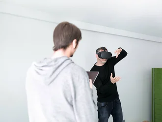 Denkströme IT-Entwickler mit VR-Brille testet virtuelle Realität im Büro der Denkströme Gruppe, betreut von Kollege mit Tablet – Symbol für innovative Digitalentwicklung und immersive Technologien