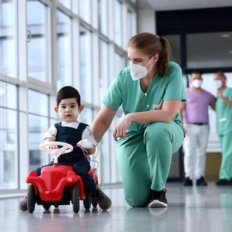 Physiotherapeutin im DHZM begleitet jungen Patienten mit Sauerstoffversorgung beim Reha-Training auf dem Spielfahrzeug – kindgerechte Therapie mit Herz