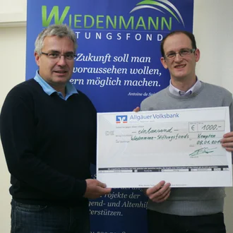 Spendenübergabe an den Wiedenmann-Stiftungsfonds 2014: Zwei Männer – einer im grauen Pullover, der andere in dunkelblauem Pullover – halten einen symbolischen Scheck der Allgäuer Volksbank über 1.000 Euro vor einem Roll-up des Wiedenmann-Stiftungsfonds