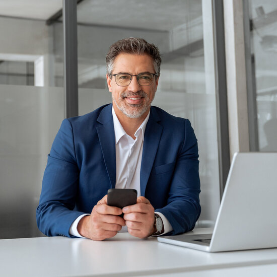 Geschäftsmann sitzt mit Smartphone und Laptop in einem modernen Büro.