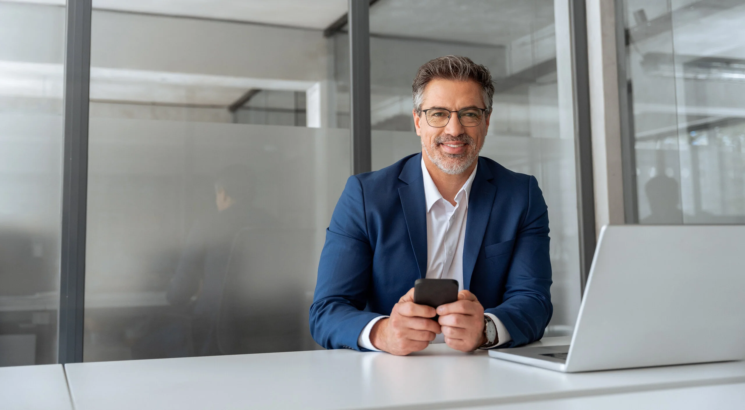 Geschäftsmann sitzt mit Smartphone und Laptop in einem modernen Büro.