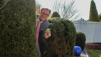 Denkströme CEO im Anzug mit pinker Krawatte blickt hinter einer Hecke hervor und hält ein bemaltes Osterei in der Hand – fröhliche Ostereiersuche im Garten bei Sonnenschein.