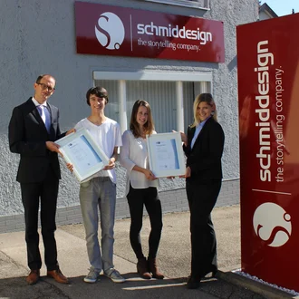 Vier Personen vor dem Agenturgebäude von schmiddesign, zwei junge Absolvent:innen erhalten feierlich ihre IHK-Zertifikate zur bestandenen Abschlussprüfung 2017
