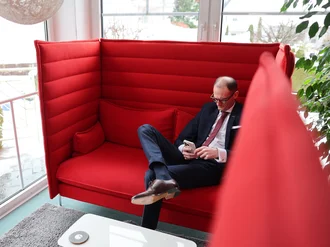 Denkströme Geschäftsführer im Anzug sitzt entspannt in moderner roter Lounge-Sitznische und nutzt Smartphone – Rückzugsort für konzentriertes Arbeiten im Büro.
