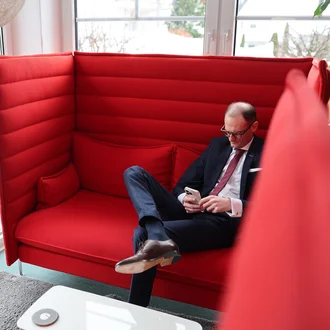 Denkströme Geschäftsführer im Anzug sitzt entspannt in moderner roter Lounge-Sitznische und nutzt Smartphone – Rückzugsort für konzentriertes Arbeiten im Büro.