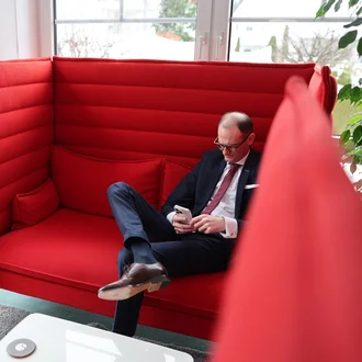 Denkströme Geschäftsführer im Anzug sitzt entspannt in moderner roter Lounge-Sitznische und nutzt Smartphone – Rückzugsort für konzentriertes Arbeiten im Büro.