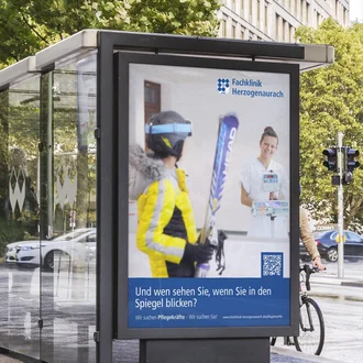 Großflächenplakat der Fachklinik Herzogenaurach mit Pflegekampagne an Bushaltestelle im urbanen Straßenverkehr vor moderner Architektur.